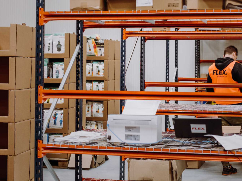 A person wearing an orange safety vest labeled "FLEX" handles a cardboard box in a warehouse. Shelves with more boxes are visible in the background. The tone is busy and industrial.